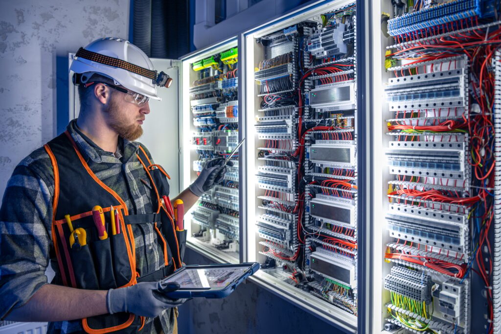 Electrician in overalls, focused on work in switchboard with fuses, using tablet.
